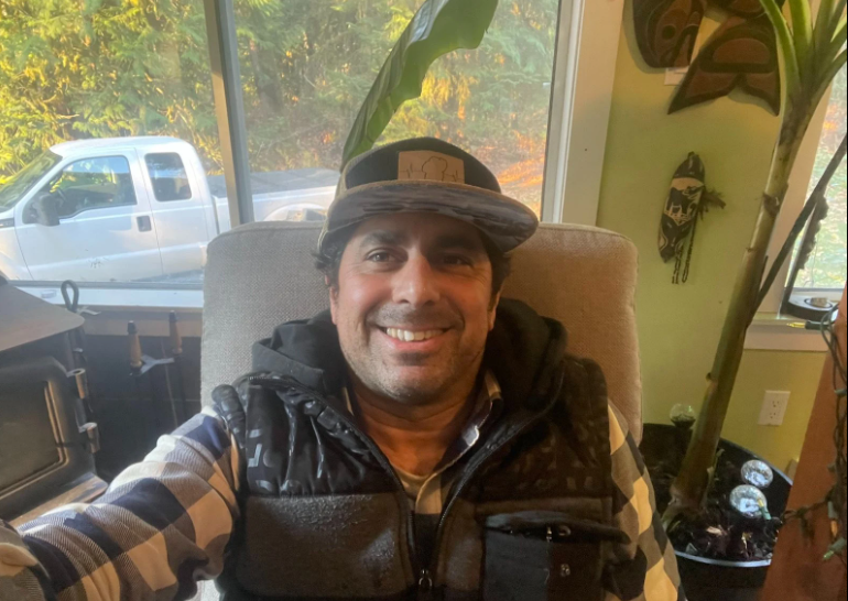 Davin grinning at the camera from his chair at home in the Squamish Valley, in a cap and puffer vest