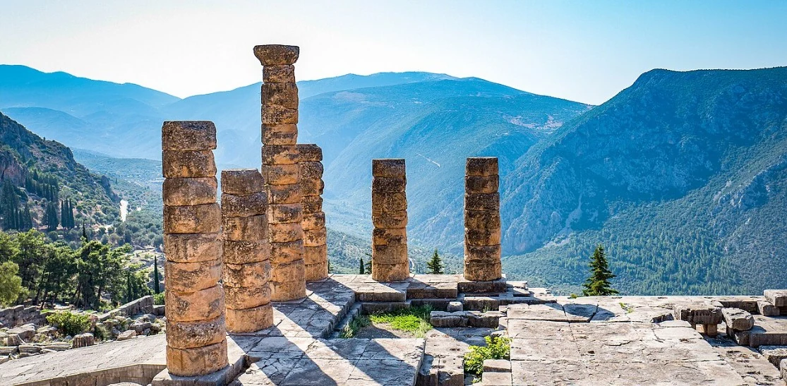 The remaining Doric columns of the Temple of Apollo at Delphi, with forested mountains and a deep valley behind them.
